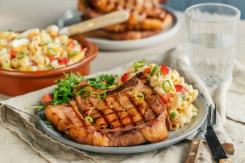 Saftige sommerkoteletter med makaronisalat servert med grønn salat på grå tallerken. Tallerkenen står på dekket middagsbord. Fat med koteletter i bakgrunnen, og en skål makaronisalat ved siden av.
