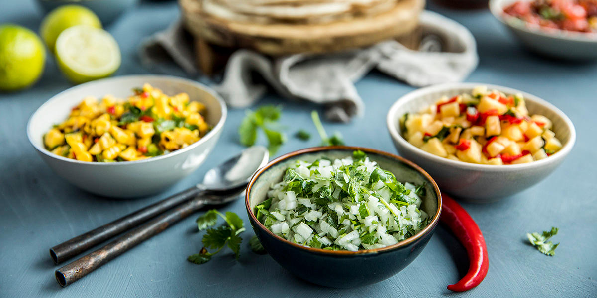 Tre skåler, i midten finhakket løk og koriander, til venstre grillet maissalsa med chili, til høyre frisk ananassalsa med chili og koriander.