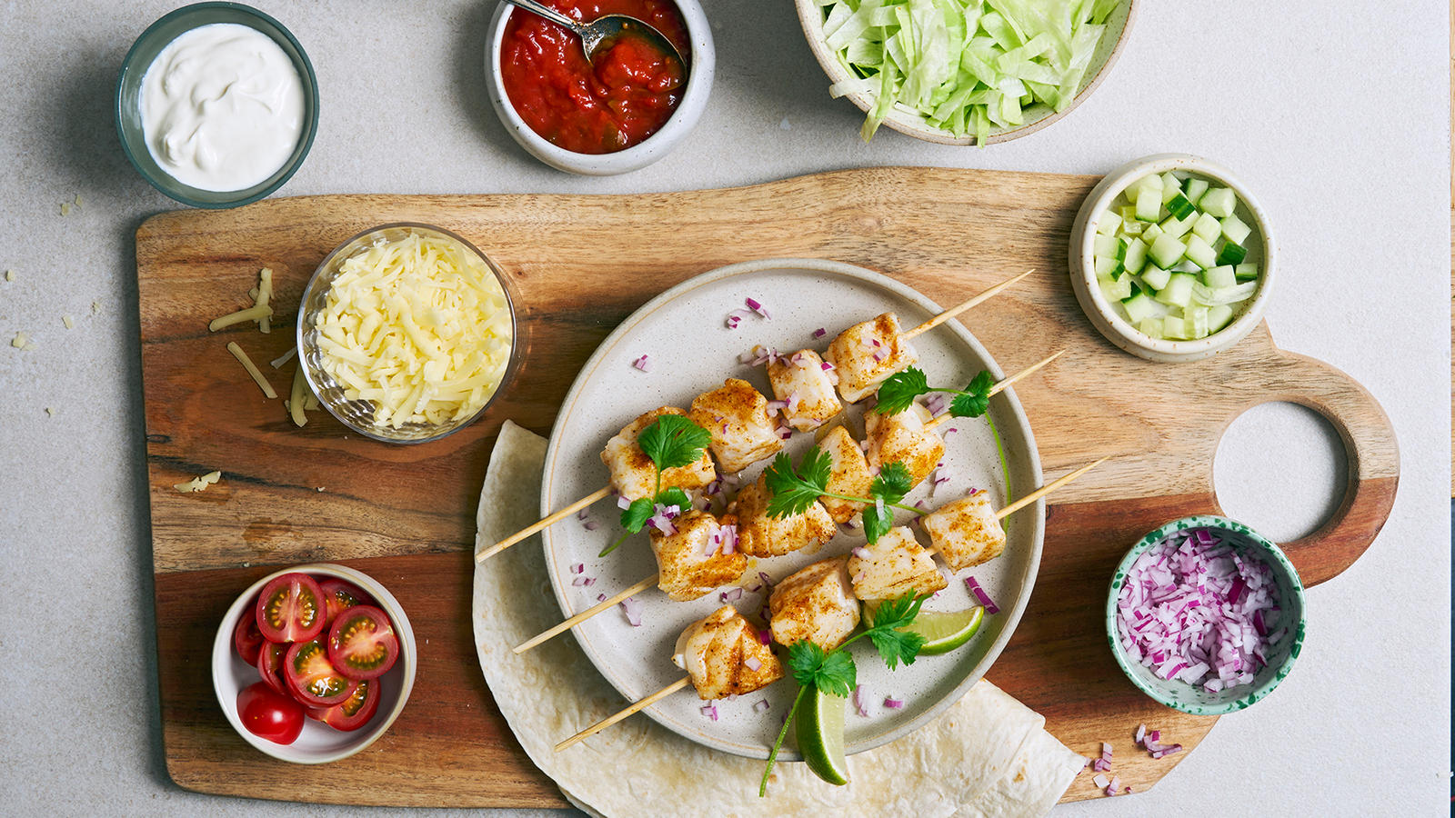 Taco med skrei servert på grå tallerken med frisk koriander og limebåter. Tacotilbehør i små boller rundt.