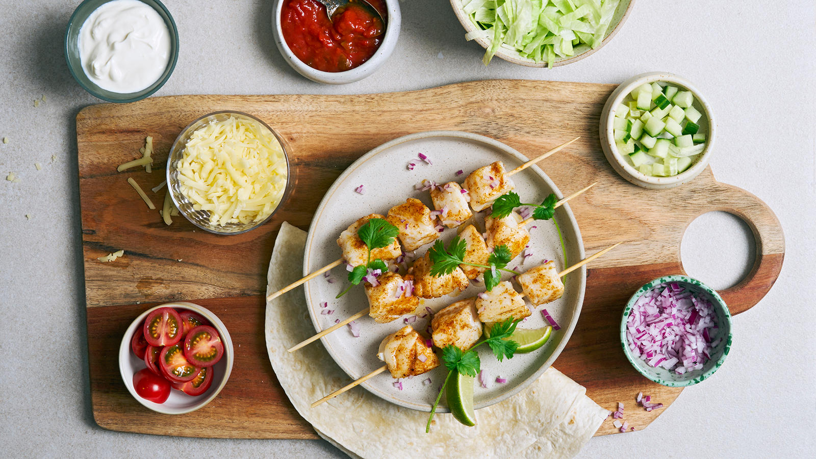 Taco med skrei servert på grå tallerken med frisk koriander og limebåter. Tacotilbehør i små boller rundt.