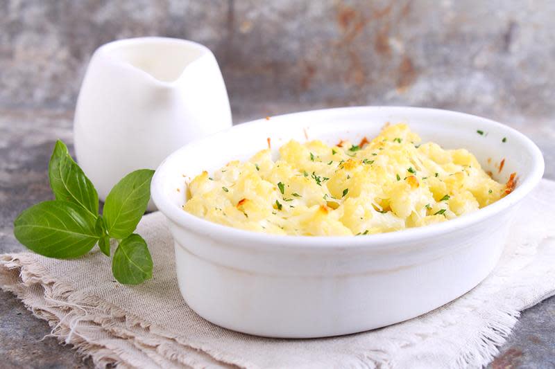 Gyllen blomkålgrateng i hvit form, servert på naturfarget linserviett, med en kvist basilikum på siden.