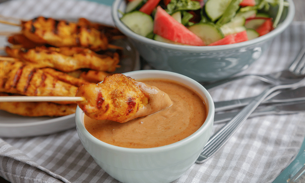 En kyllingbit på grillspyd blir dyppet i peanøttsaus. I bakgrunnen ligger det flere kyllingspyd på en tallerken og en bolle med salat.