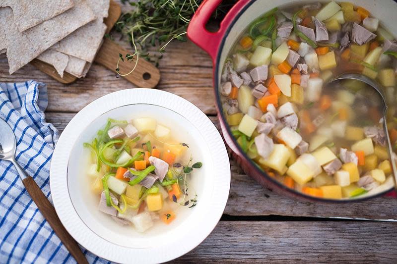 Fargerik betasuppe i rød støpejernskjele og hvit suppeskål, servert på rustikk trebord med blårutet kjøkkenhåndkle. Flatbrød og ferske urter ved siden av.