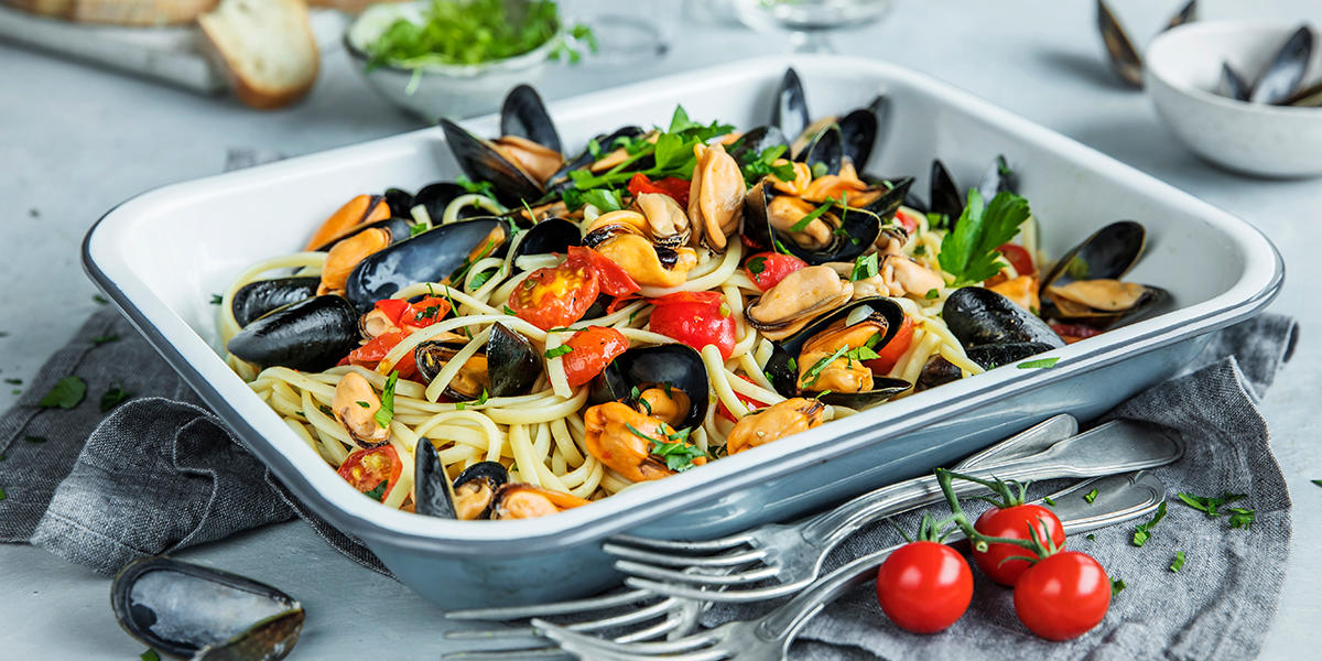 Linguine med blåskjell og eplemost servert i ildfast form med tomater og friske urter, på dekket middagsbord. Godt brød, ferske urter og blåskjellskall i bakgrunnen.