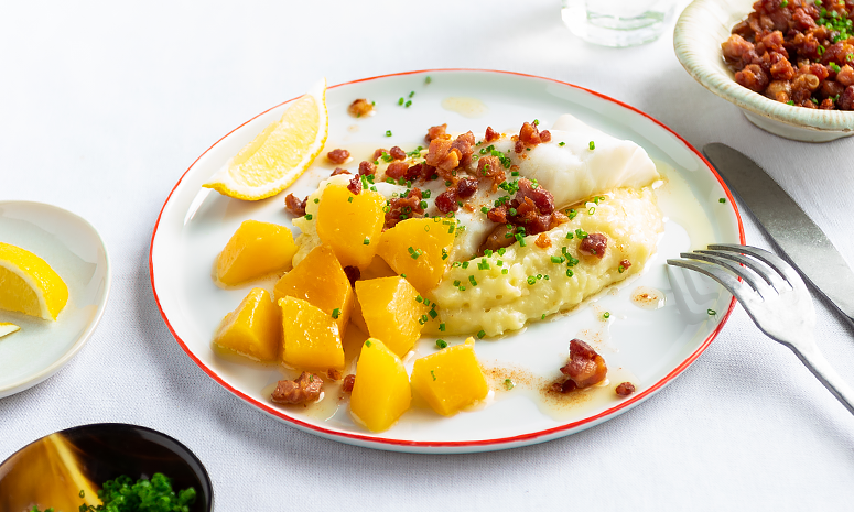 Lettsaltet torsk, potetmos og kålrot pyntet med gressløk, bacon og en sitronbåt på hvit tallerken med rød kant.