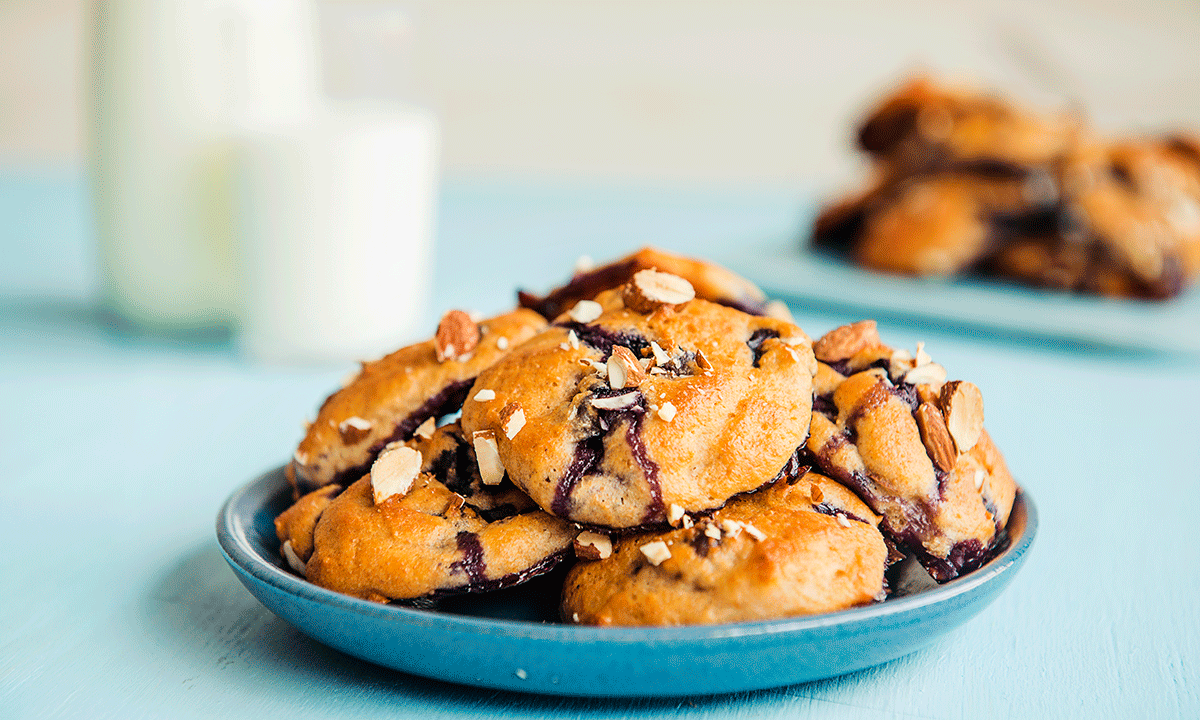 Yoghurtkjeks med blåbær servert på blå asjett på blått bord med melkeglass i bakgrunnen. Kjeksene er pyntet med skivede mandler.