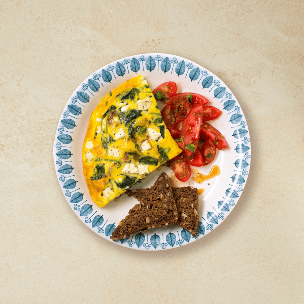 Et stykke ovnsbakt omelett med feta, spinat og tomatsalat servert på hvit blåmønstrert tallerken på beige bakgrunn, med to stykker ristet brød.
