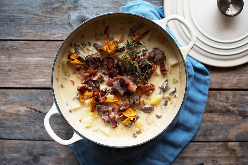 Stor hvit støpejernsgryte på rustikk trebakgrunn. Gryten er fylt med finnbiff og toppet med bacon, kantareller og fersk timian.