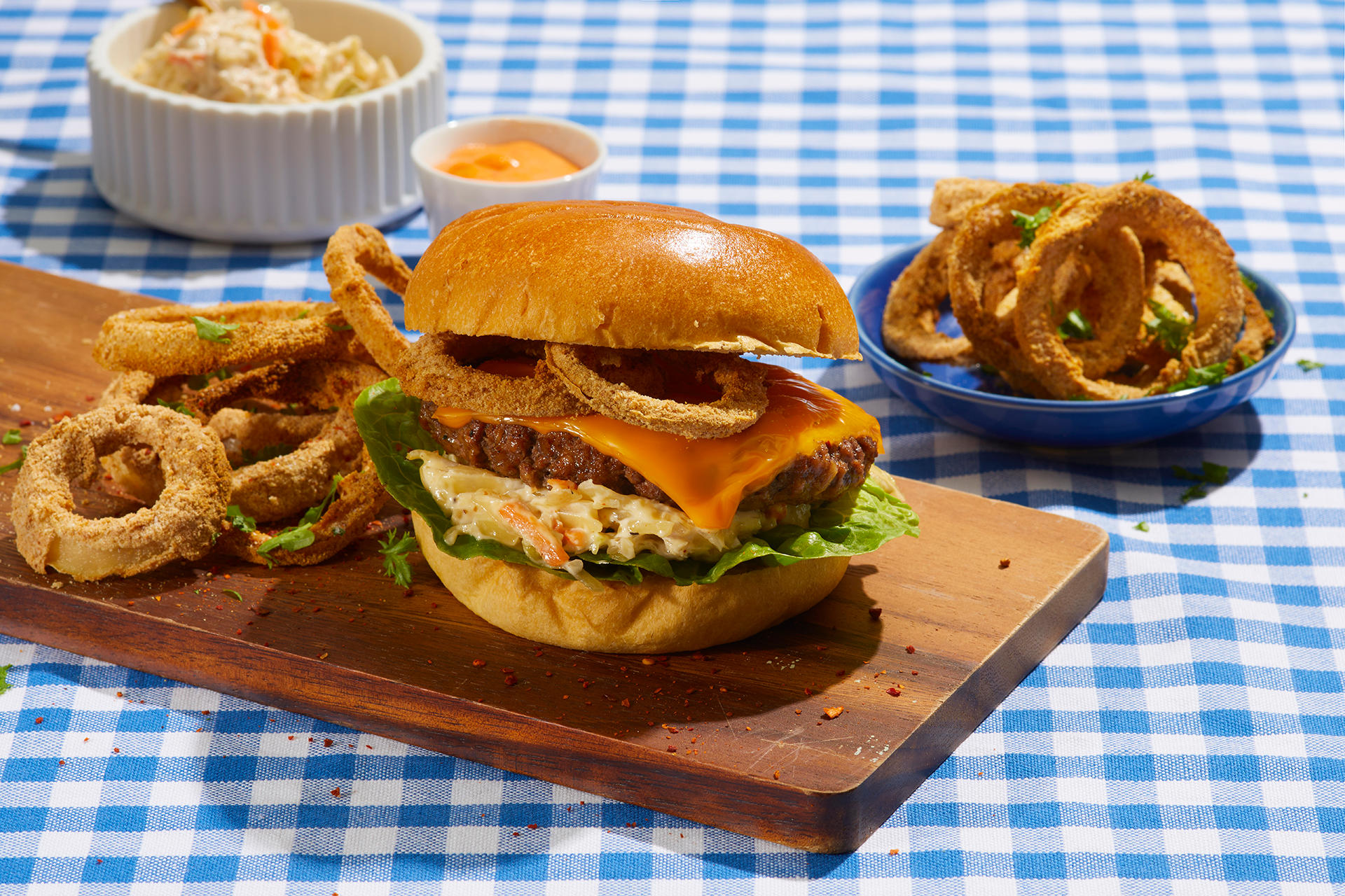 Chili burger med hjemmelagede løkringer og srirachadip, servert i brioche burgerbrød med coleslaw, cheddar og salatblad, på trefjøl på blårutete duk. Blå skål med løkringer, og hvit skål med coleslaw i bakgrunnen.