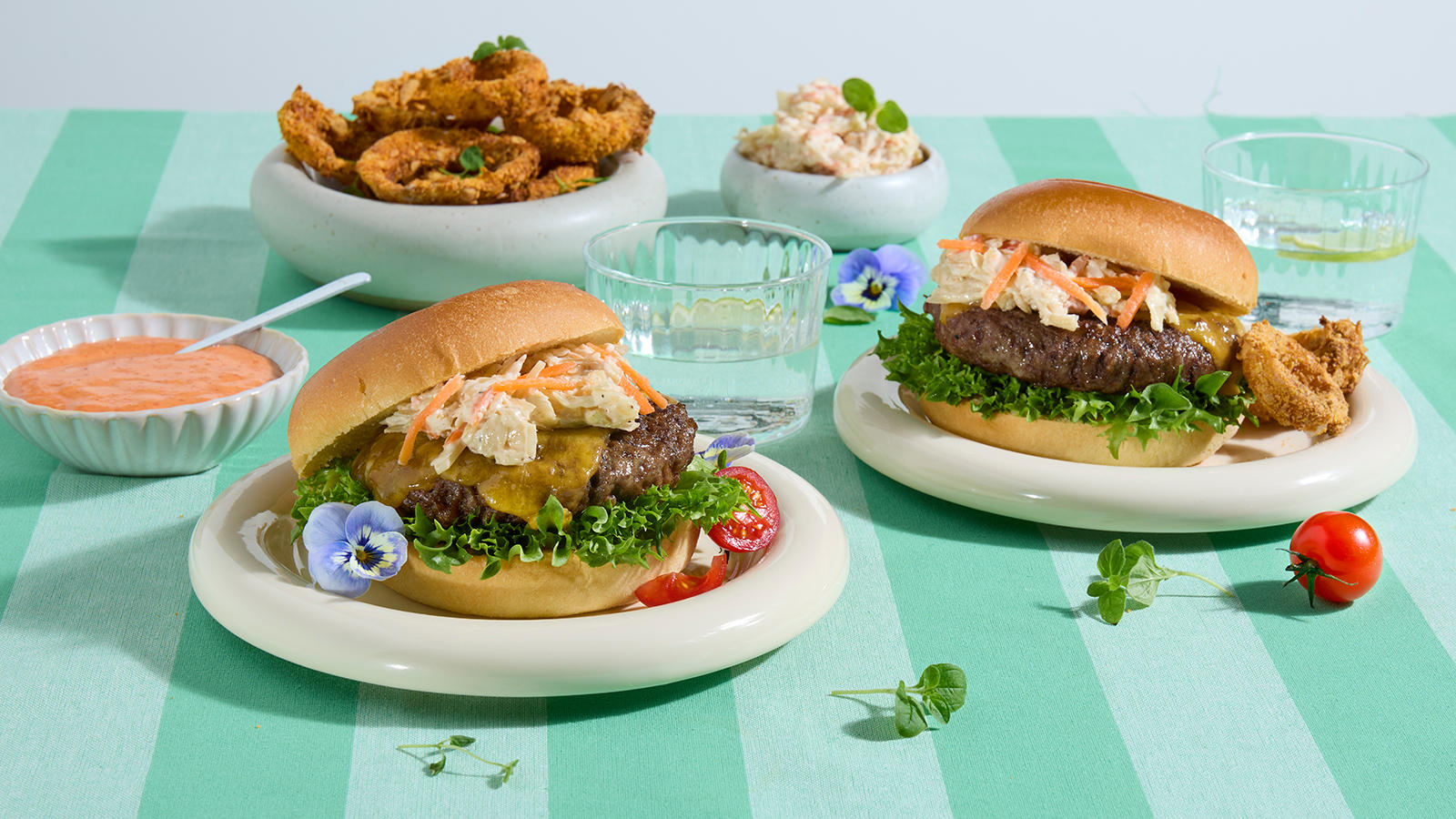 Burger med hjemmelagede løkringer og srirachadip servert på lyse asjetter på grønn stripete duk. Skåler med srirachadip og hjemmelagede løkringer i bakgrunnen.