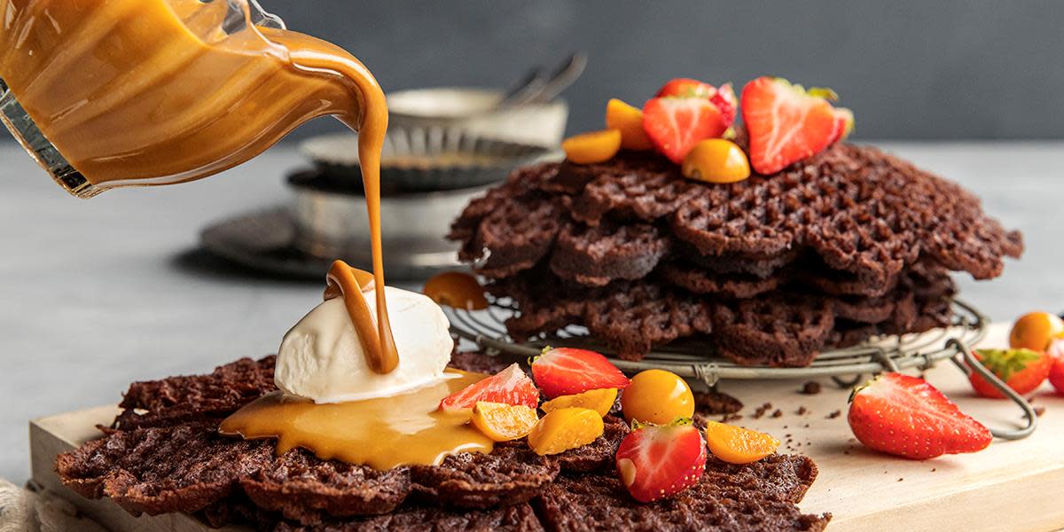 Glutenfrie sjokoladevafler med salt karamellsaus, servert med iskrem, ferske jordbær og physalis. Stabel med sjokoladevafler i bakgrunnen, og en mugge med salt karamellsaus som helles over vaflene foran.