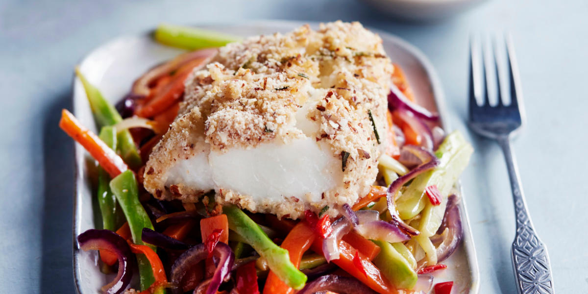 Mandelpanert skrei servert på en seng av grønnsaker på hvit firkantet tallerken med metallgaffel ved siden av.