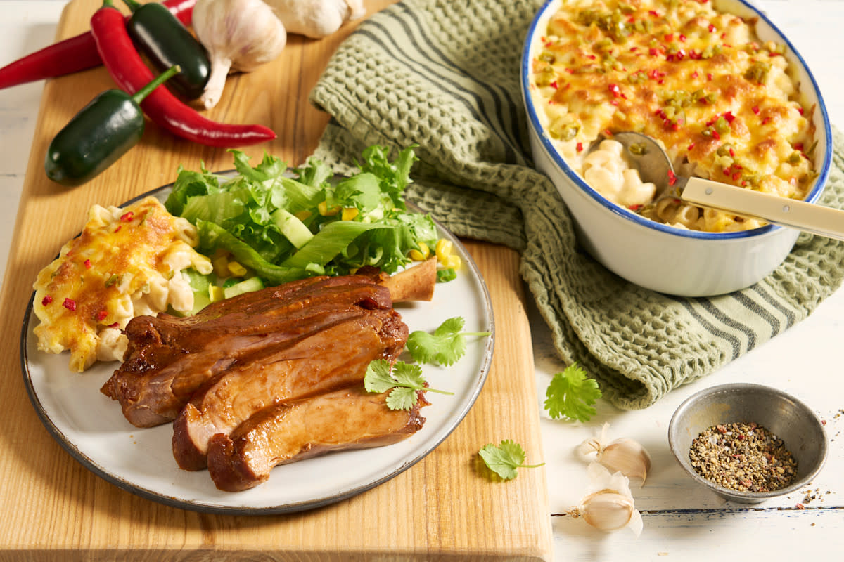Pig wings med jalapeño mac’n cheese servert på hvit tallerken på lys trefjøl med grønn salat. Frisk chili, jalapeño og hvitløk i bakgrunnen, og en form jalapeño mac'n cheese på siden.