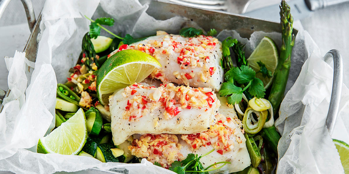 Fiskepakke på grillen med asiatiske smaker, servert i ildfast form med hvitt bakepapir, asparges, vårløk og limebåter. Fiskestykkene er dekket med smeltet hjemmelaget kryddersmør med chili og hvitløk.