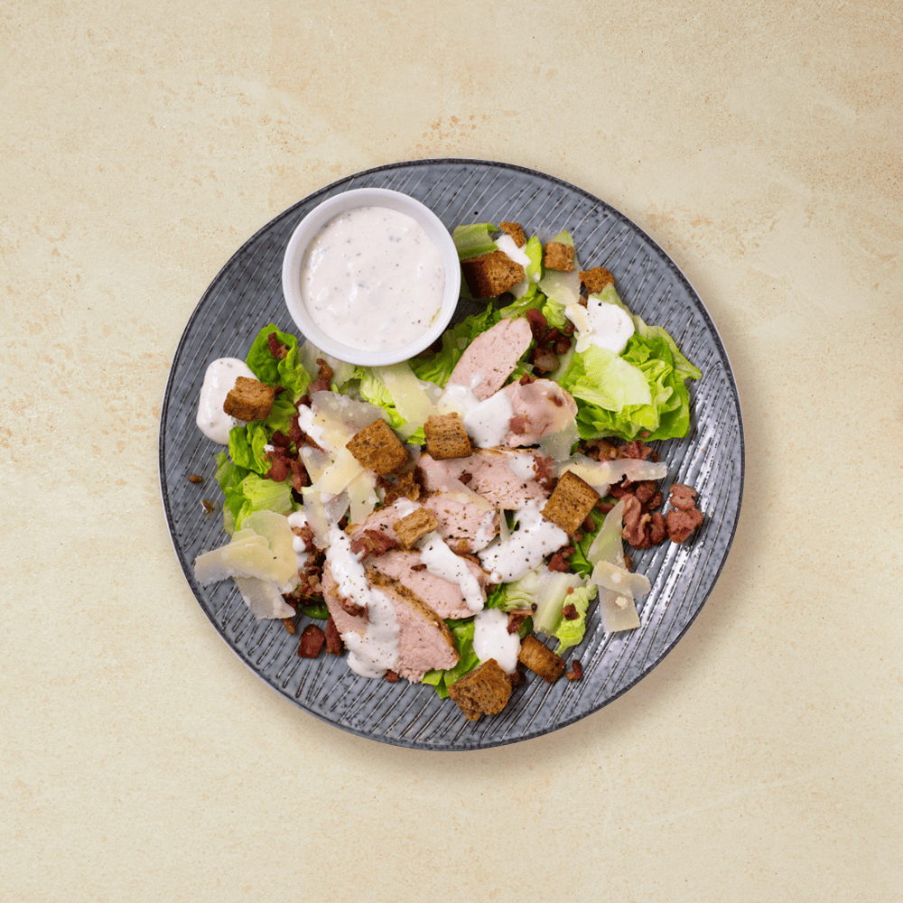 Cæsarsalat med kylling, toppet med revet parmesan og krutonger på en grå tallerken med en liten hvit skål rømmedressing.