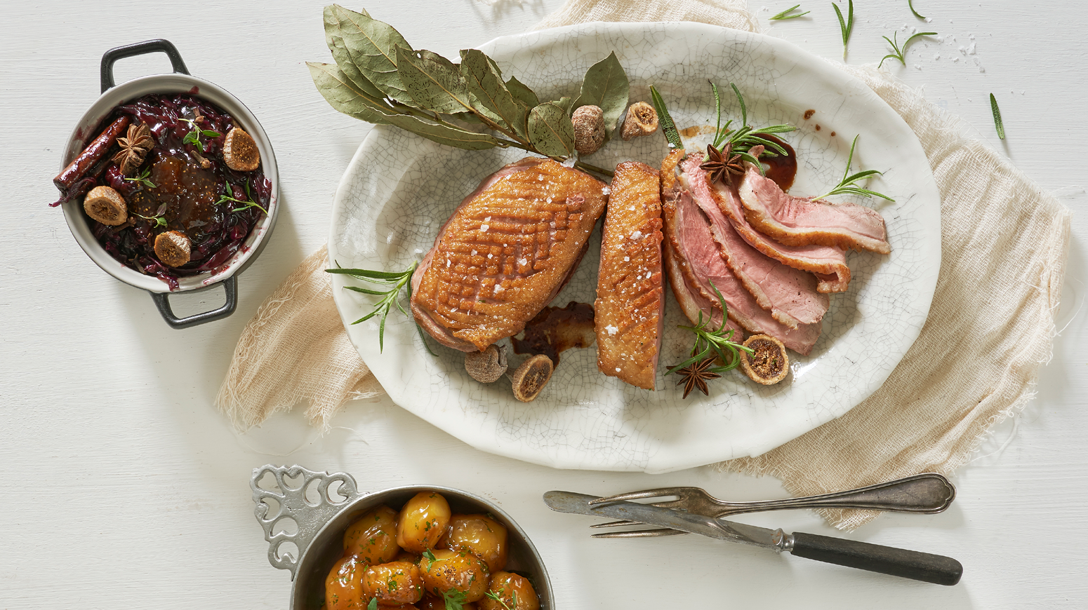 Sprøstekt andebryst servert på hvitt ovalt fat med tørkede og friske urter, og tørket fiken. Skåler med dansk rødkål og karamelliserte poteter ved siden av.