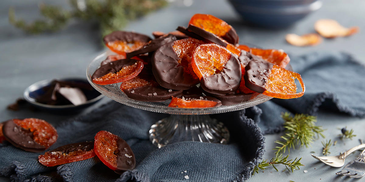 Kandiserte klementiner med sjokoladetrekk, servert på stettfat i glass. Mørkegrått kjøkkenhåndkle og grå bakgrunn.