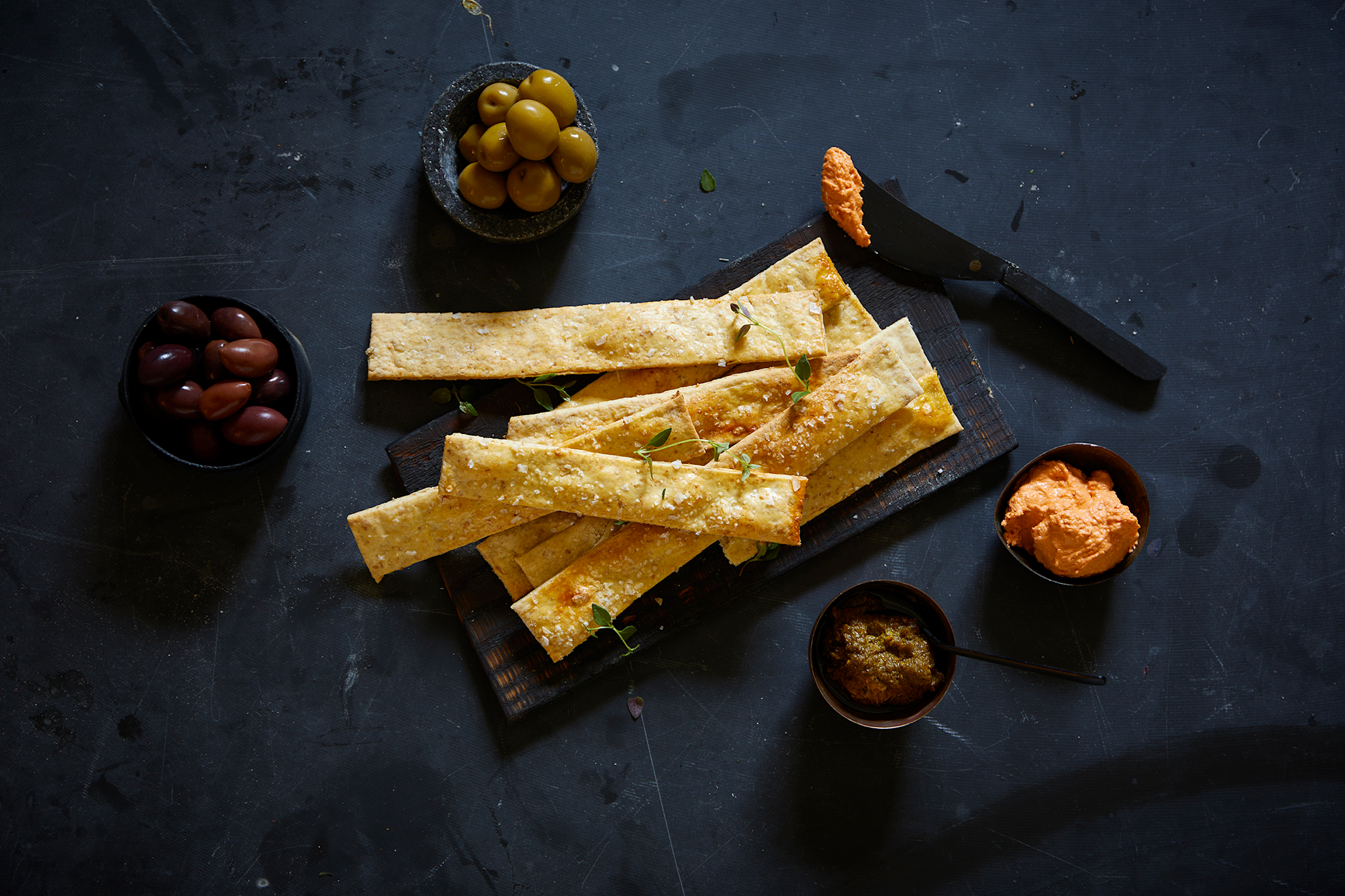 Hjemmelagede ostekjeks servert på mørk trefjøl med små skåler tilbehør rundt. I skålene ligger det blant annet oliven.