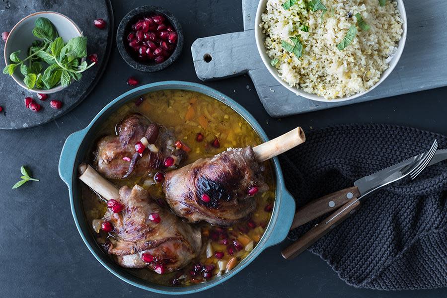 Marokkanske lammeskanker i blå gryte på mørkt bord, overstrødd med granateplefrø. Skål med couscous ved siden av, og små skåler med granateplefrø og ferske urter.