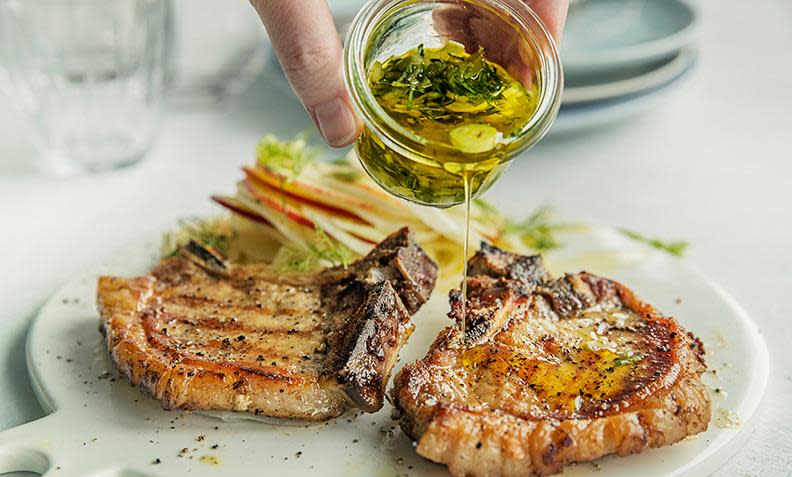 Svinekotelett med eple- og fennikelsalat servert på hvit fjøl. Urteolje blir helt overkotelettene av en hånd som holder et lite glass urteolje.