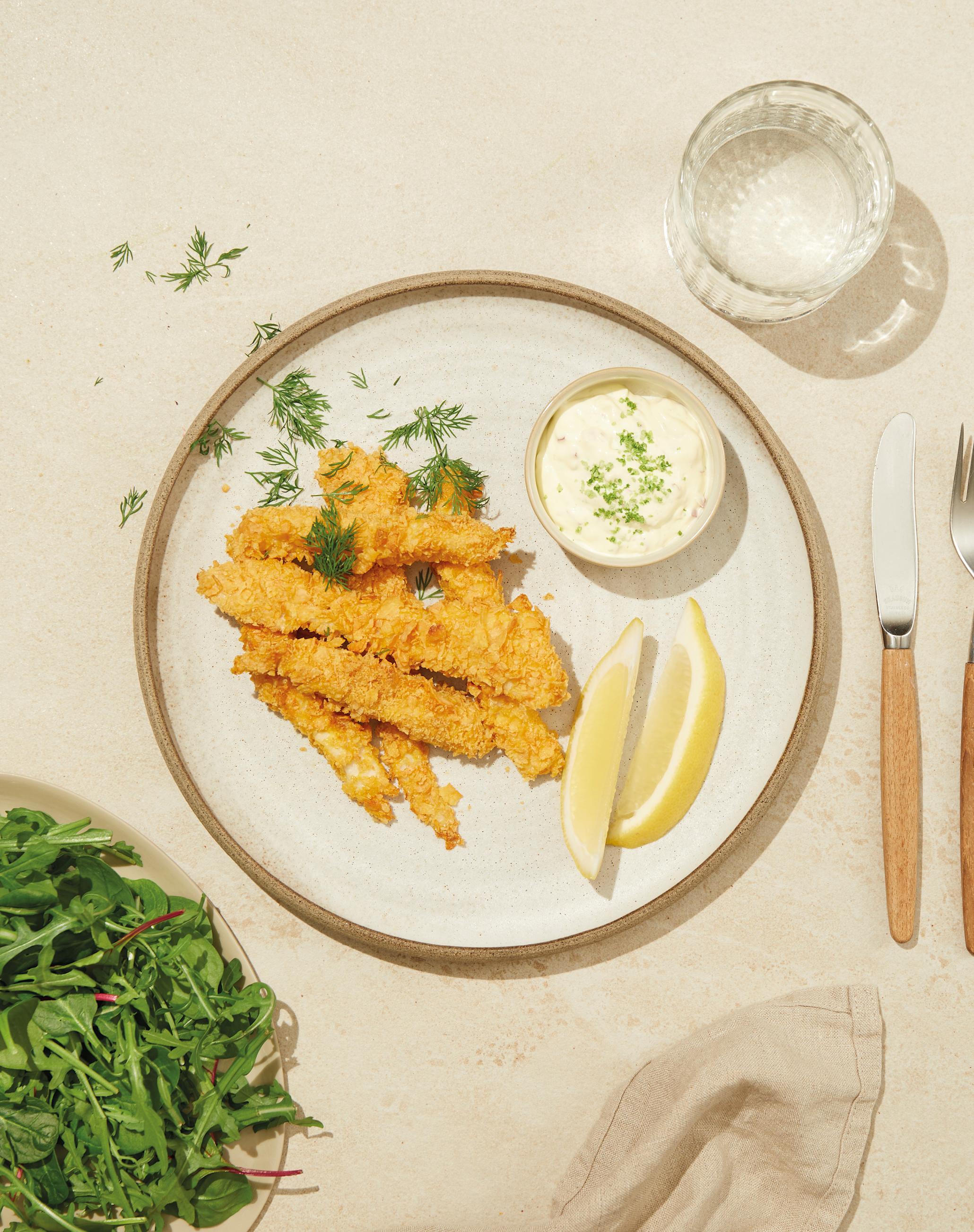 Hjemmelagde fiskepinner servert på lys tallerken med liten lys skål med hjemmelaget remulade ved siden av. Frisk dill og sitronbåter på samme tallerken. Stor skål med salatblanding ved siden av.