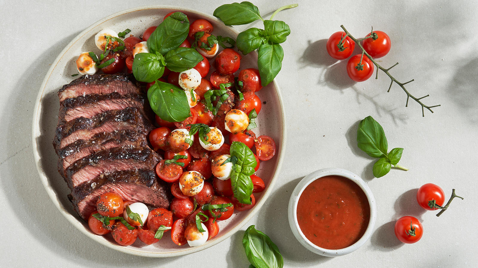 Grillet entrecôte med capresesalat og tomatdressing servert på lys keramikkasjett på lys bakgrunn. Friske piccolotomater og liten skål med tomatdressing på siden.
