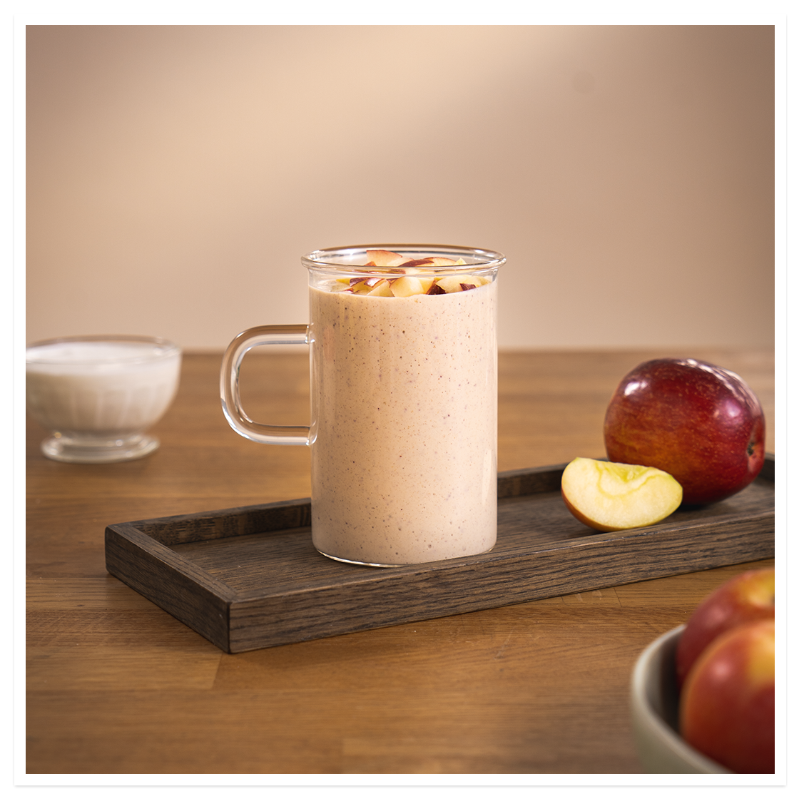 Sommersnø -smoothie, lys beige smoothie med rødt eple og peanøttsmør i glasskopp på mørk trefjøl. Rødt eple ved siden av.