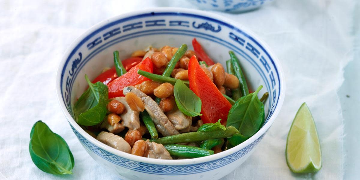 Wok med svinekjøtt og peanøtter i en blå og hvitmønstret skål, servert med aspargesbønner, paprika og fersk basilikum. En båt lime på siden.
