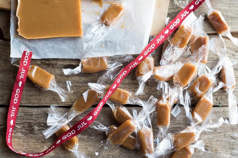 Hjemmelagede fløtekarameller pakket inn i cellofan, ligger på rustikk trebakgrunn med rødt julebånd over.