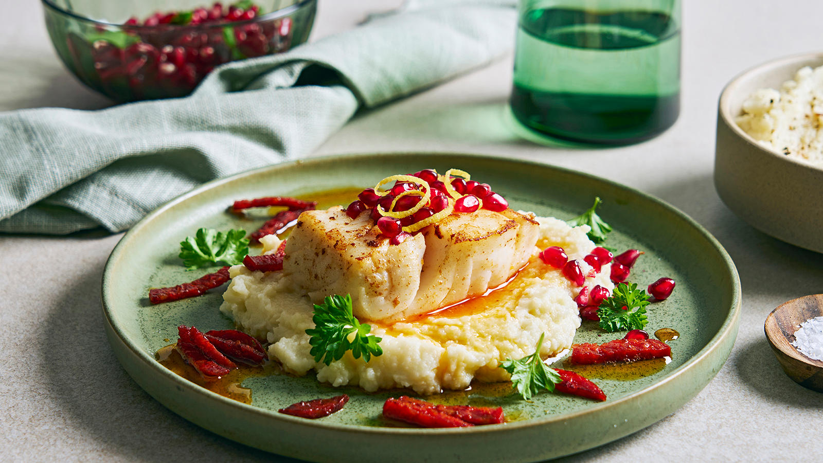 Skrei med chorizo og mandelpotetpuré servert med grønne urter og revet sitronskall på grønn tallerken på dekket middagsbord. Grønn skål med granateplefrø i bakgrunnen.