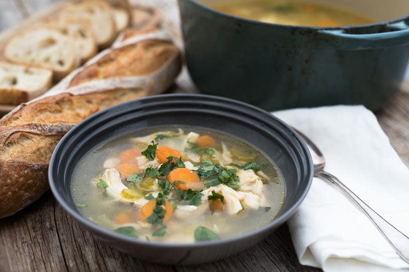 Klar kyllingsuppe med grønnsaker, toppet med persille. Servert i mørk skål, rustikk baguett og lys linserviett ved siden av.