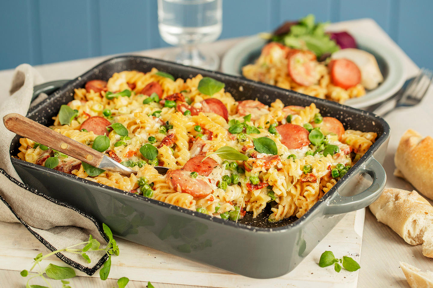 Pølsegrateng med pasta, pølser, soltørket tomat, erter og ost servert i mørk grå ildfast form på lys trefjøl. Formen står på dekket middagsbord.