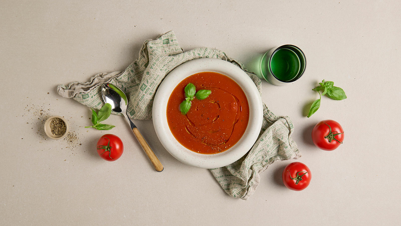 Ovnsbakt tomatsuppe servert i lys skål med olivenolje, frisk basilikum og nykvernet pepper på toppen. Friske tomater og basilikum rundt.