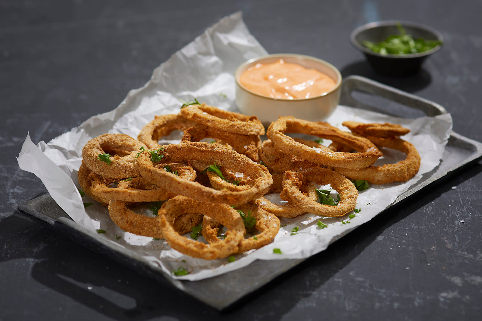Hjemmelagede løkringer med spicy srirachadip servert med et dryss grønne urter på hvitt bakepapir på mørkt metallfat. Hvit skål med srirachadip på fatet med løkringene, og liten skål med grønne urter i bakgrunnen.