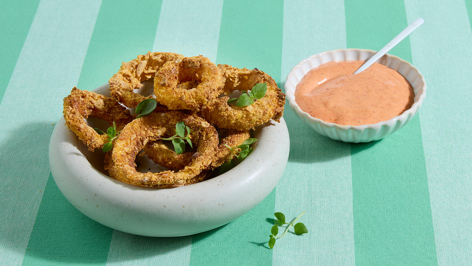 Hjemmelagede løkringer med spicy srirachadip servert i lys skål med et dryss grønne urter. Liten skål srirachadip på siden.
