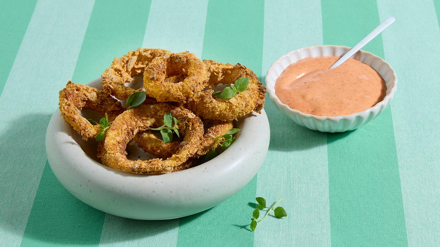 Hjemmelagede løkringer med spicy srirachadip servert i lys skål med et dryss grønne urter. Liten skål srirachadip på siden.