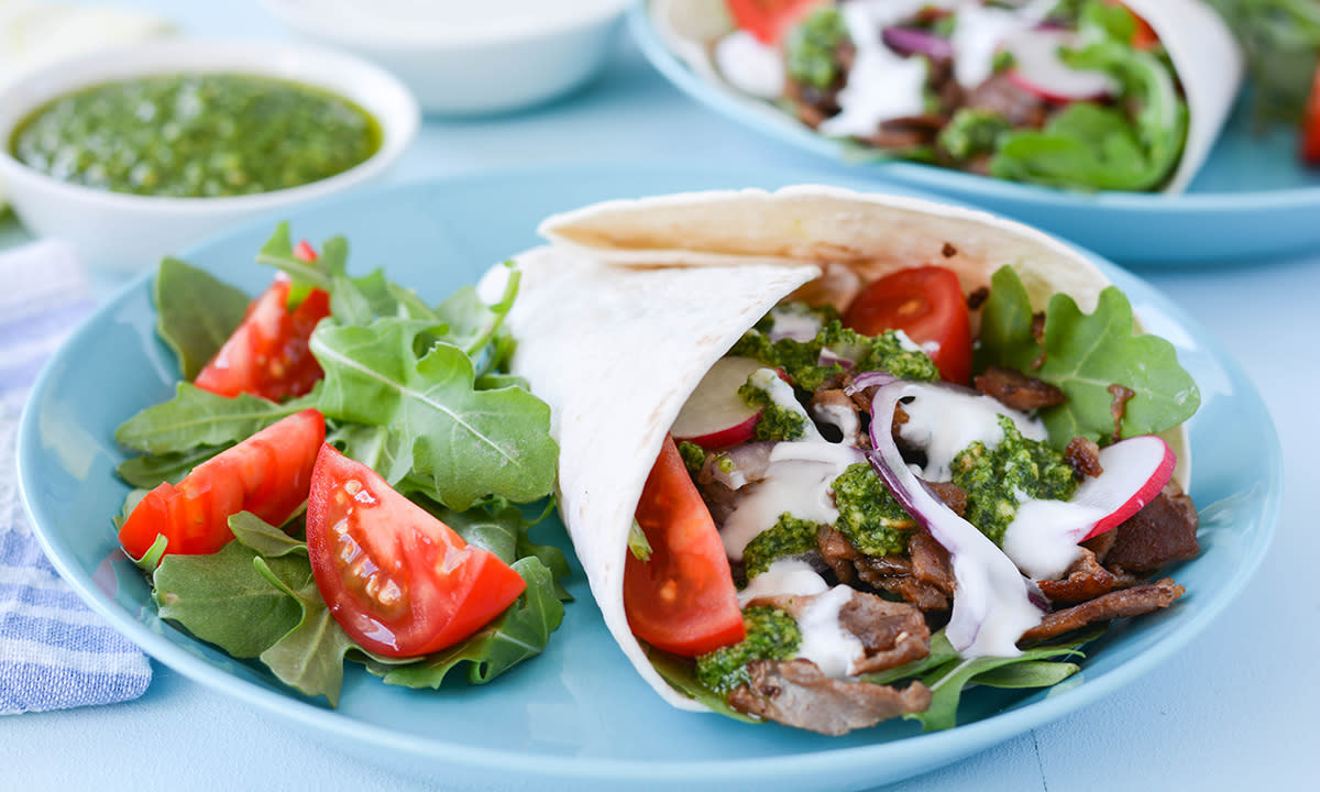 Kebab med lam, pesto, grønnsaker og dressing servert i tortillalefse på lys blå tallerken. Skål med pesto i bakgrunnen.
