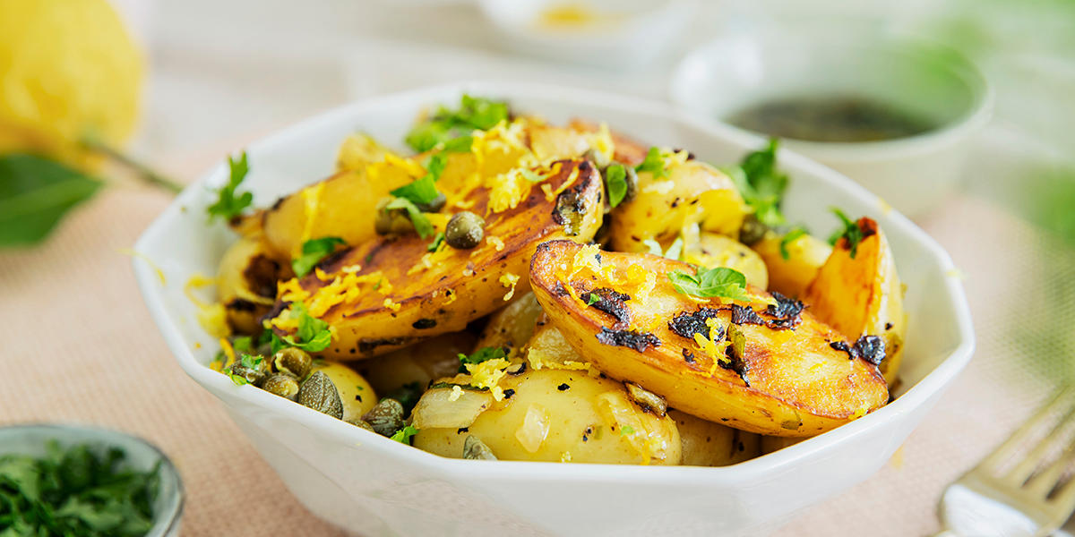 Grillet potetsalat med sitron og kapers, servert i hvit skål med sitronskall og ferske urter drysset over. Fersk hel sitron i bakgrunnen og en liten skål urter ved siden av.