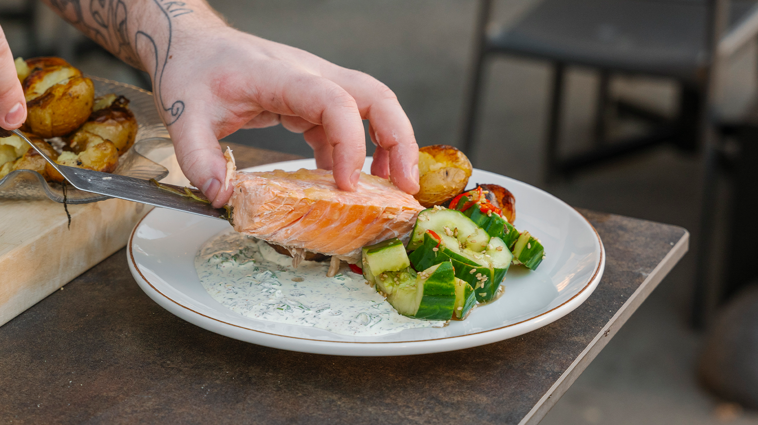 Laks i folie med agurksalat, småpoteter og rognsaus servet uten folien med stekespade over på hvit tallerken på kanten av et restaurantbord.