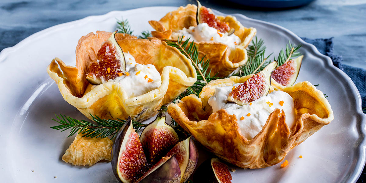 Hjemmelagde krumkaker formet til skåler, servert med ovnsbakte fiken og marsipankrem med et dryss finrevet klementinskall på hvit tallerken. Desserten er pyntet med små granbar.