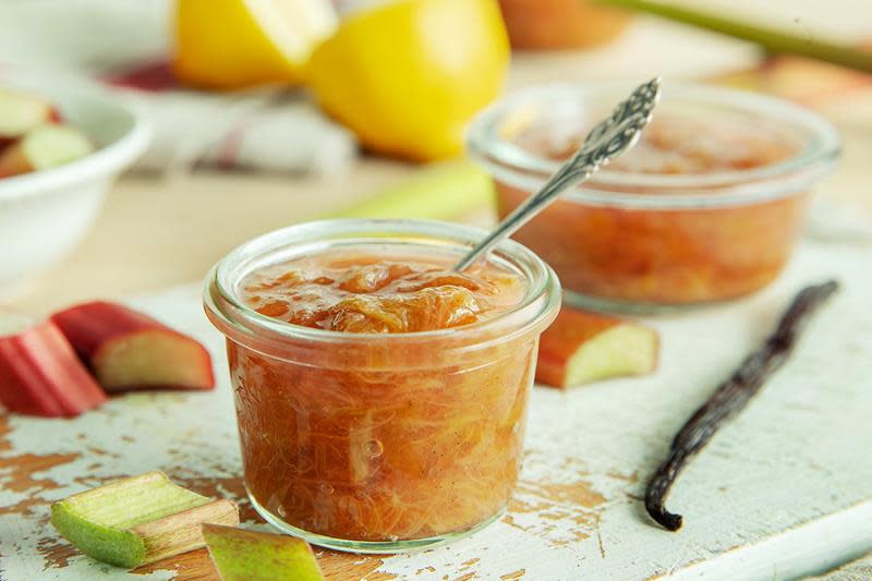 Lyst rødlig rabarbrasyltetøy på gammeldags syltetøyglass, med sølvskje. Servert på rustikk malt treplate, vaniljestang og sitron på siden.