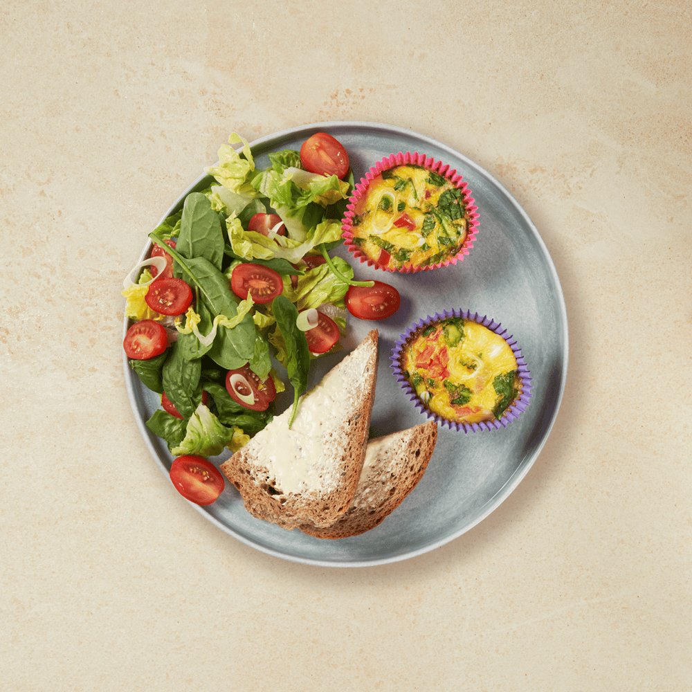 To stykk spinatfrittati i muffinsformer servert på grå tallerken på beige bakgrunn, med grønn salat og brødskiver med smør.
