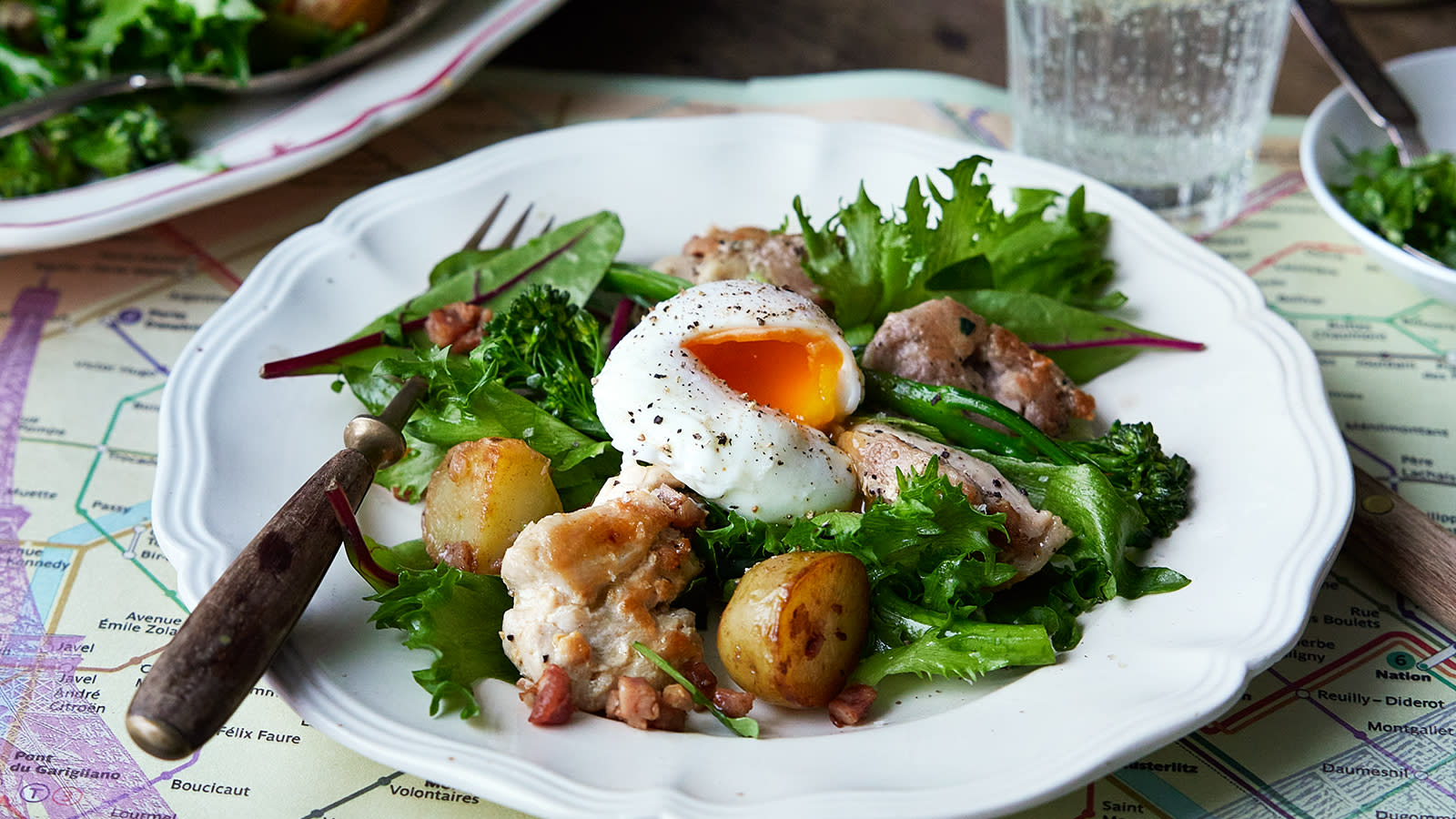 Salade lyonnaise med kylling – kyllingsalat med lune posjerte egg servert på hvit tallerken på dekket middagsbord, med rustikk gaffel på tallerkenen.