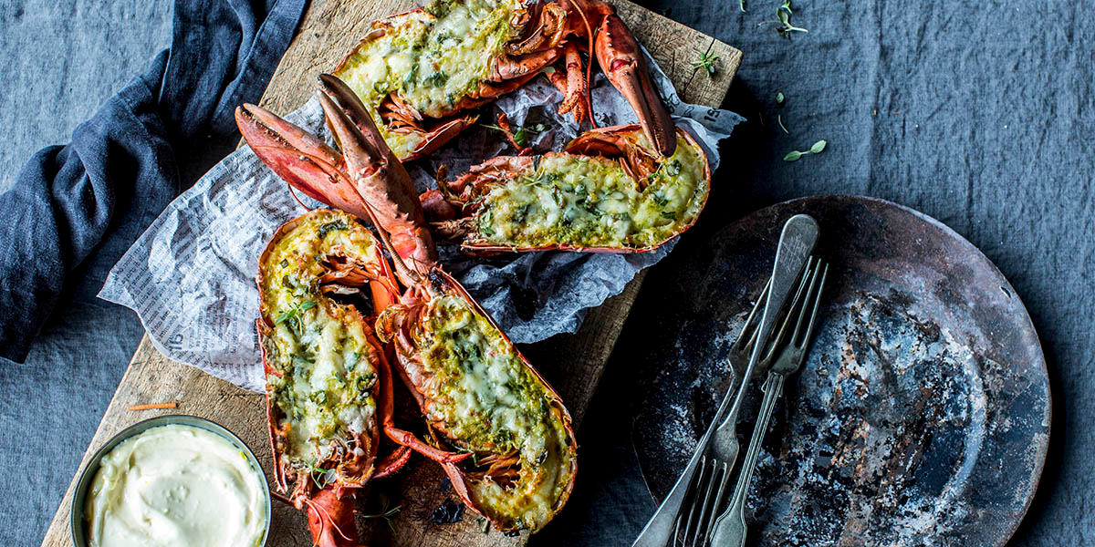 Gratinert hummer med Gruyère, delt i to og servert på rustikk trefjøl på mørk linduk. Liten skål med aioli ved siden av.