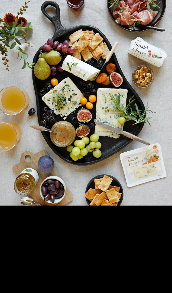 Et utvalg Smak* oster servert på organisk svart trefat med druer, fiken, bjørnebær, pærer, tørket aprikos og Smak* kjeks. Forskjellige oster i pack rundt fatet.