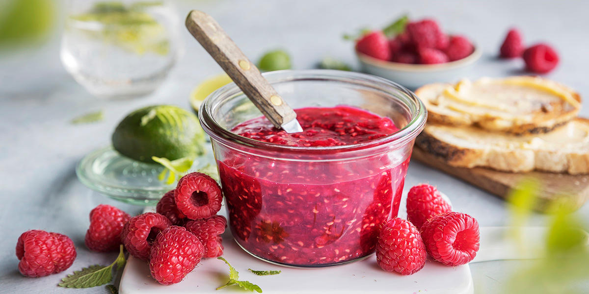 Glass med bringebærsyltetøy med skje i. Rundt ligger friske bringebær, en halv lime og to loffskiver med smør.