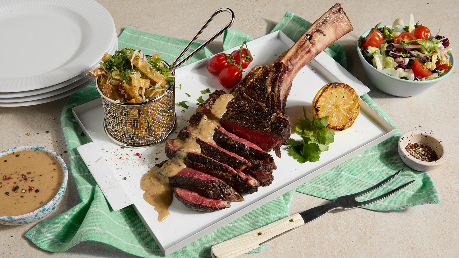 Tomahawk biff med hjemmelaget peppersaus servert på hvitt fat med parmesan fries, grillet sitron og cherrytomater. Skål med peppersaus ved siden av.