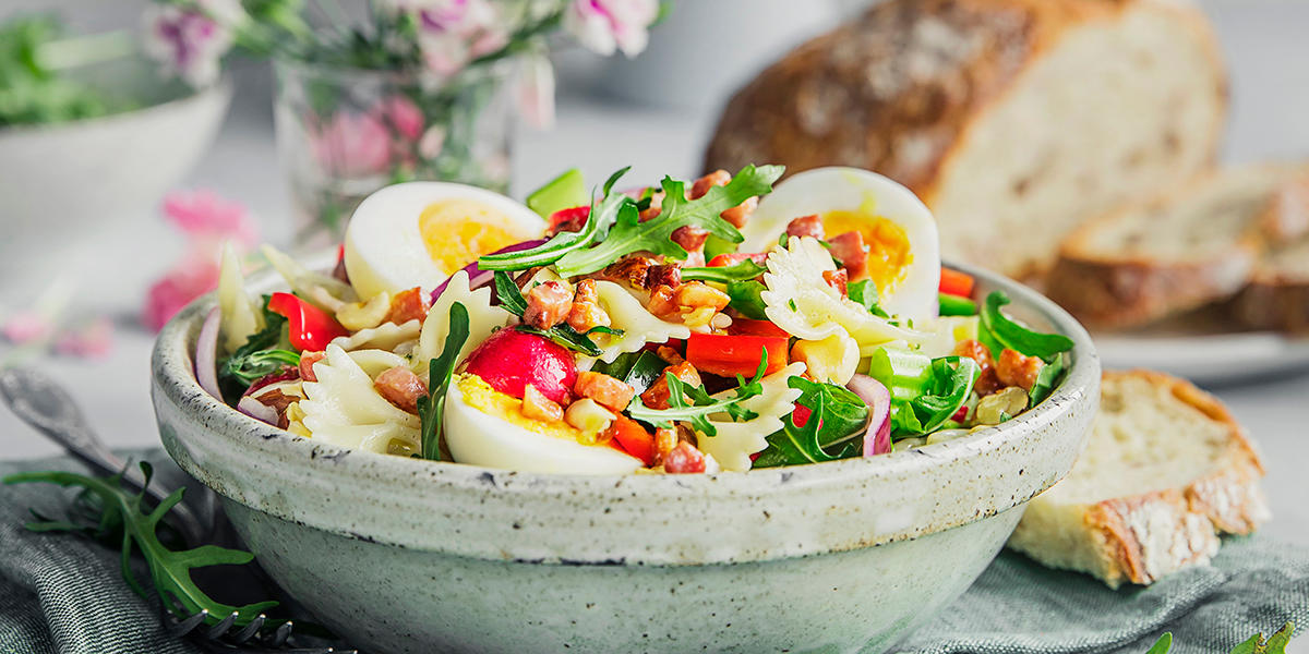 Fargerik og mettende pastasalat med egg, bacon, pasta, ruccola og grønnsaker, servert i lys bolle med landbrød til.