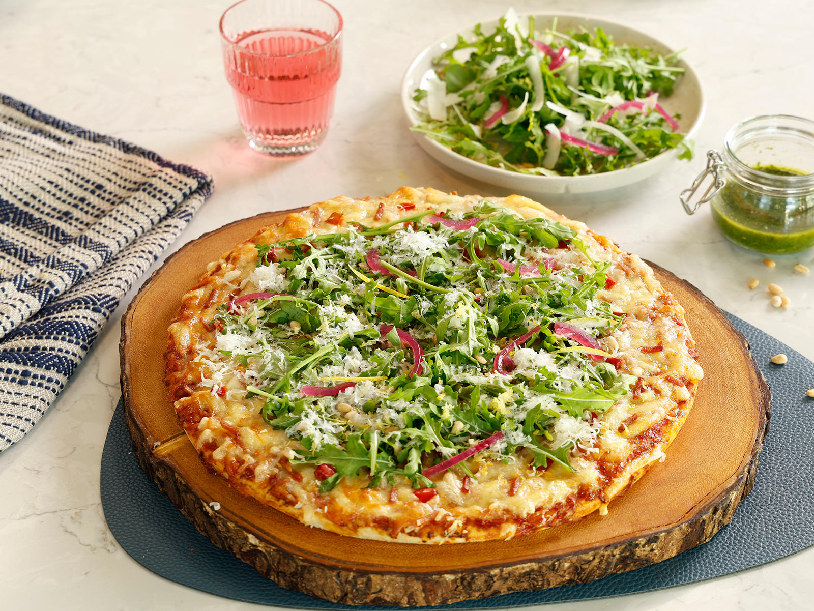 Salattopping med parmesan til pizza Grandiosa servert på en nystekt Grandiosa Original på rund rustikk trefjøl. Glass med saft, skål med salat og glass med grønn pesto i bakgrunnen.