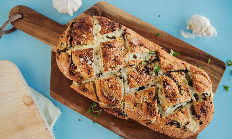 Hvitløksbrød med mozzarella og urtesmør, laget av et helt landbrød. Servert på trefjøl, toppet med ferske urter. Blå bakgrunn.